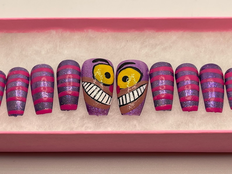 K&ouml;nnte beinhalten: Ein Set aus k&uuml;nstlichen Fingern&auml;geln mit einer lila Glitzerbasis und rosa Streifen sowie Designs der Grinsekatze. Die Grinsekatzen-N&auml;gel haben eine lila Basis, gelbe Augen und ein breites Grinsen. Die N&auml;gel werden in einer rosa Schachtel pr&auml;sentiert.