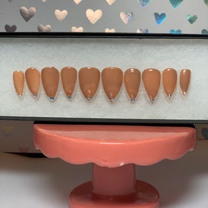 May include: A set of almond-shaped, press-on nails in a nude color, each with a small, iridescent gem at the base. The nails are arranged in a black box, displayed on a pink, tiered stand against a background with heart patterns.