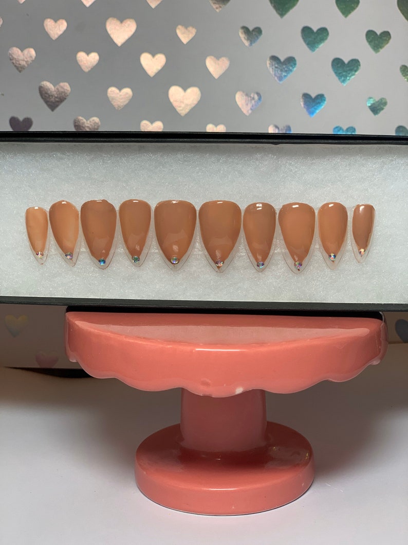 May include: A set of almond-shaped, press-on nails in a nude color, each with a small, iridescent gem at the base. The nails are arranged in a black box, displayed on a pink, tiered stand against a background with heart patterns.