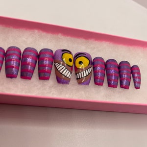 K&ouml;nnte beinhalten: Ein Set aus zehn aufklebbaren N&auml;geln in einer rosa Schachtel. Die N&auml;gel haben eine lila Glitzerbasis mit rosa Streifen und zwei N&auml;gel mit einem Cheshire Cat-Design. Die Cheshire Cat-N&auml;gel haben ein braunes Gesicht, gelbe Augen und ein breites Grinsen.