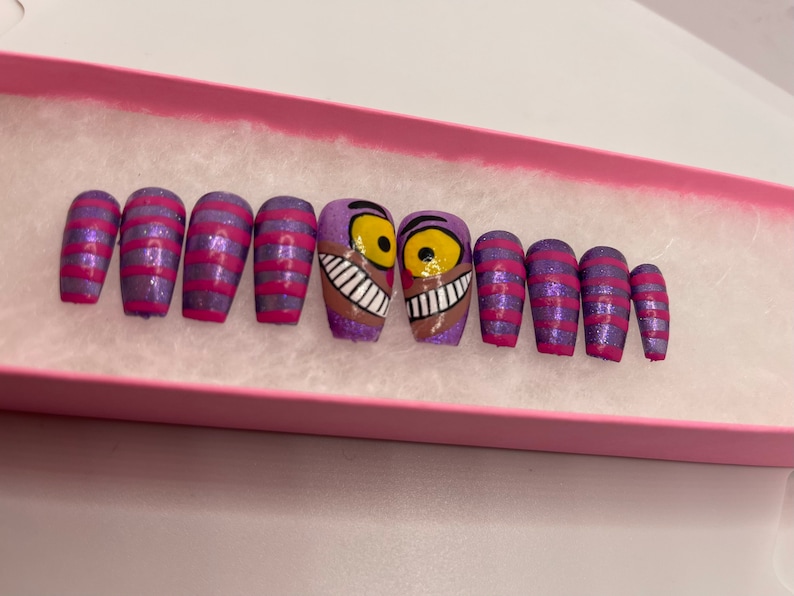 K&ouml;nnte beinhalten: Ein Set aus zehn aufklebbaren N&auml;geln in einer rosa Schachtel. Die N&auml;gel haben eine lila Glitzerbasis mit rosa Streifen und zwei N&auml;gel mit einem Cheshire Cat-Design. Die Cheshire Cat-N&auml;gel haben ein braunes Gesicht, gelbe Augen und ein breites Grinsen.