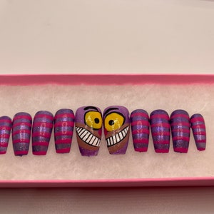 K&ouml;nnte beinhalten: Ein Set von aufklebbaren N&auml;geln mit einem lila-rosa gestreiften Design und Glitzer. Zwei N&auml;gel zeigen ein Cartoon-Gesicht der Grinsekatze mit gelben Augen und einem breiten Grinsen. Die N&auml;gel werden in einer rosa Box pr&auml;sentiert.