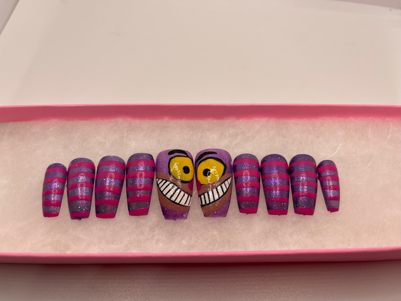 K&ouml;nnte beinhalten: Ein Set von aufklebbaren N&auml;geln mit einem lila-rosa gestreiften Design und Glitzer. Zwei N&auml;gel zeigen ein Cartoon-Gesicht der Grinsekatze mit gelben Augen und einem breiten Grinsen. Die N&auml;gel werden in einer rosa Box pr&auml;sentiert.