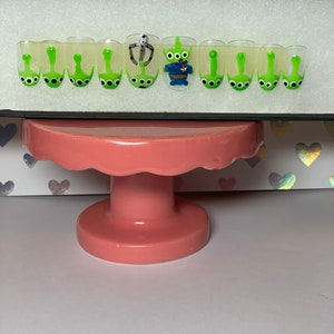 May include: A set of press-on nails featuring a Toy Story alien theme. Each nail is clear with a green alien design, and one nail has a 3D alien figure. The nails are displayed on a pink cake stand.