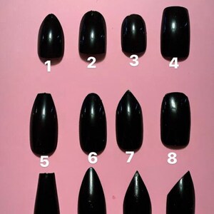 May include: A collection of twelve glossy black press-on nails in various shapes, including oval, almond, and square, arranged on a pink background. Each nail is numbered for easy selection.