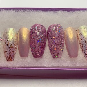 May include: A set of press-on nails in a variety of designs. Some nails are a shimmering pink, others are covered in glitter, and some have iridescent accents. Several nails feature decorative rhinestones. The nails are displayed in a purple box.