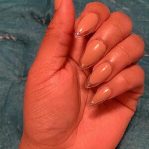 May include: A hand with almond-shaped nails painted in a nude color with a clear, glittery border. The nails are long and pointed, with small, iridescent gems near the cuticle. The skin tone is medium brown.
