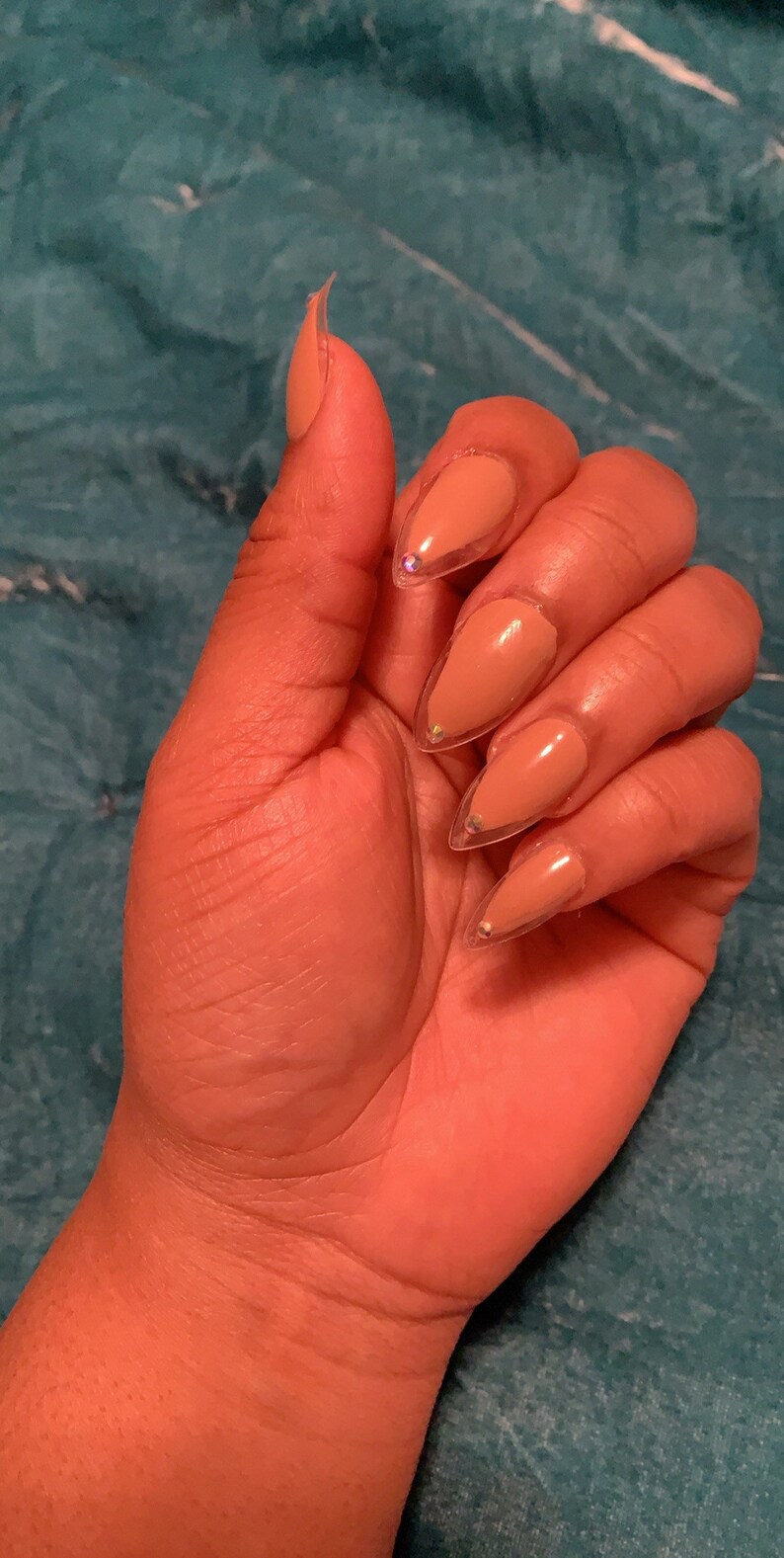 May include: A hand with almond-shaped nails painted in a nude color with a clear, glittery border. The nails are long and pointed, with small, iridescent gems near the cuticle. The skin tone is medium brown.