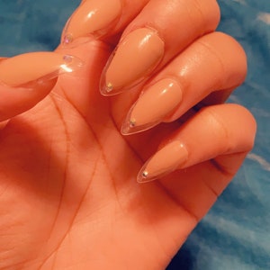 May include: Close-up of a hand with long, almond-shaped acrylic nails. The nails are painted a nude color with clear tips and small, sparkling rhinestones at the base of each nail. The hand is resting on a blue surface.