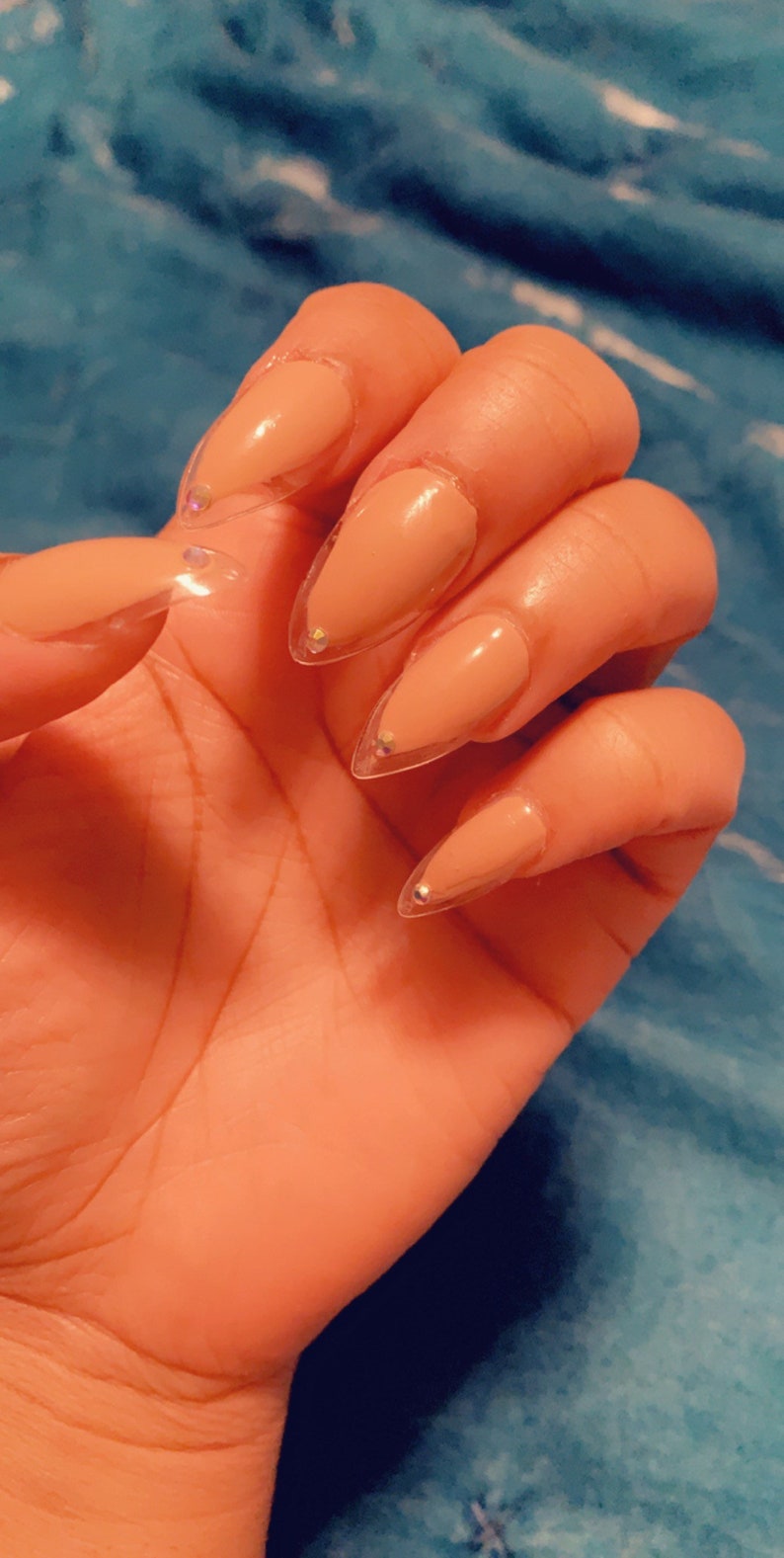 May include: Close-up of a hand with long, almond-shaped acrylic nails. The nails are painted a nude color with clear tips and small, sparkling rhinestones at the base of each nail. The hand is resting on a blue surface.