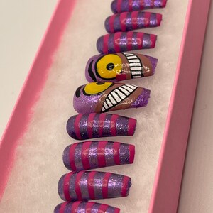 K&ouml;nnte beinhalten: Ein Set k&uuml;nstlicher N&auml;gel in einer rosa Schachtel. Die N&auml;gel sind lila mit rosa Glitzer und rosa Streifen. Ein Nagel zeigt ein Design der Grinsekatze mit einem gelben Auge und einem grinsenden Mund.