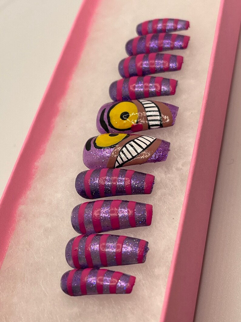 K&ouml;nnte beinhalten: Ein Set k&uuml;nstlicher N&auml;gel in einer rosa Schachtel. Die N&auml;gel sind lila mit rosa Glitzer und rosa Streifen. Ein Nagel zeigt ein Design der Grinsekatze mit einem gelben Auge und einem grinsenden Mund.