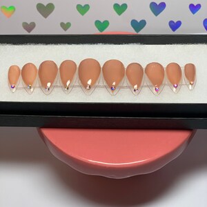 May include: A set of ten almond-shaped, nude-colored press-on nails. Each nail features a small, iridescent gem near the cuticle. The nails are arranged in a black box, presented on a pink surface with a background of holographic heart shapes.