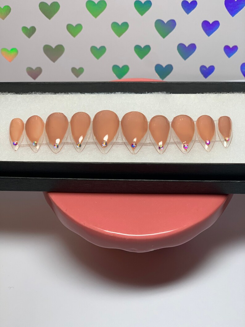 May include: A set of ten almond-shaped, nude-colored press-on nails. Each nail features a small, iridescent gem near the cuticle. The nails are arranged in a black box, presented on a pink surface with a background of holographic heart shapes.