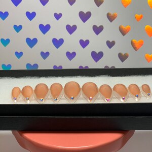 May include: A set of ten artificial nails in a black box. The nails are a nude color with a clear, glittery tip and a small, iridescent gem at the base. The background features a pattern of iridescent hearts.
