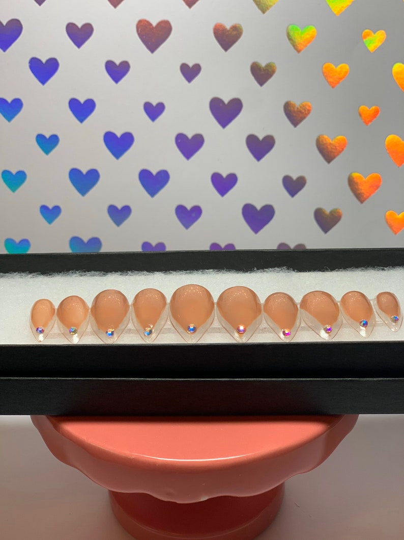 May include: A set of ten artificial nails in a black box. The nails are a nude color with a clear, glittery tip and a small, iridescent gem at the base. The background features a pattern of iridescent hearts.