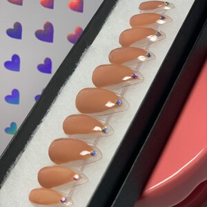 May include: A set of ten almond-shaped false nails in a neutral beige color, each with a clear tip and a small iridescent gem. The nails are arranged in a black box with a white interior.