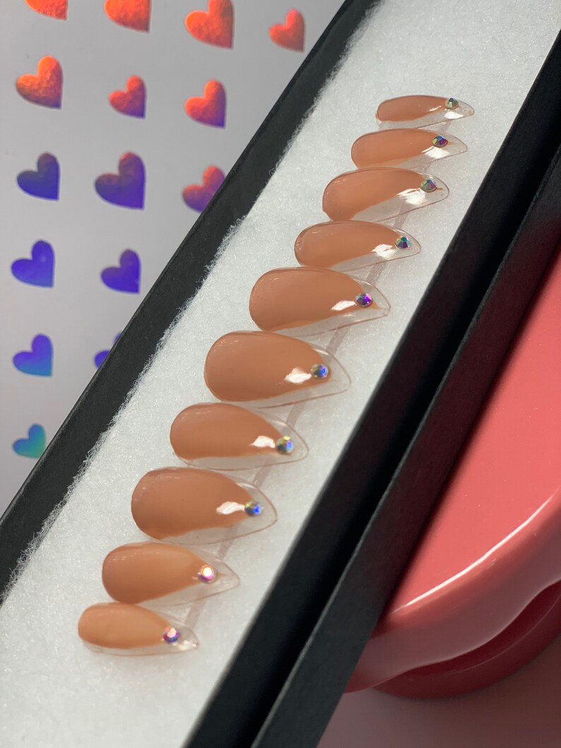 May include: A set of ten almond-shaped false nails in a neutral beige color, each with a clear tip and a small iridescent gem. The nails are arranged in a black box with a white interior.