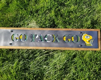 Puede incluir: Un letrero de madera con una placa de metal que dice "GO PACK GO" en letras verdes y amarillas. La placa de metal está rodeada por un marco de madera. Las letras están recortadas de la placa de metal y el fondo es un patrón de tela verde y amarillo. Un contorno del estado de Wisconsin en verde y amarillo con un casco de fútbol está en el lado derecho del letrero.