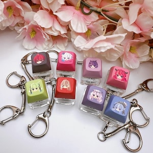 May include: A collection of keychains featuring colorful keycaps with anime-style character illustrations. Each keycap is a different color, including red, pink, green, and purple. The keychains have silver metal hardware and are displayed against a backdrop of pink flowers.