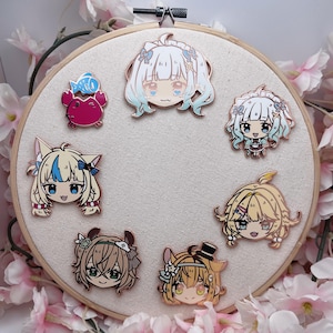 May include: A collection of seven anime-style enamel pins, featuring various character designs with detailed facial features and colorful accents. The pins are arranged on a white fabric stretched within a wooden embroidery hoop, surrounded by pink flowers.
