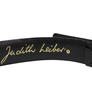 K&ouml;nnte beinhalten: Nahaufnahme eines schwarzen Lederg&uuml;rtels mit goldener Schnalle. Die Worte "Judith Leiber" sind in goldener Schrift auf der Innenseite des G&uuml;rtels geschrieben.