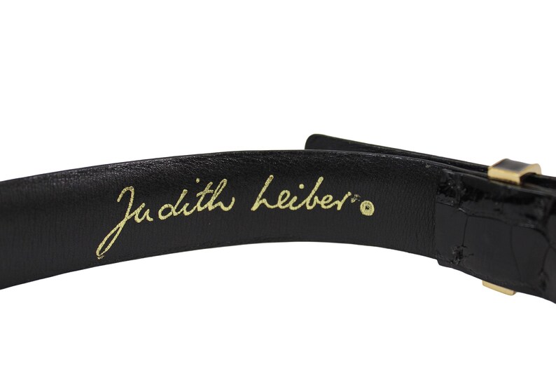 K&ouml;nnte beinhalten: Nahaufnahme eines schwarzen Lederg&uuml;rtels mit goldener Schnalle. Die Worte "Judith Leiber" sind in goldener Schrift auf der Innenseite des G&uuml;rtels geschrieben.