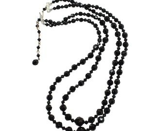 May include: A three-strand black beaded necklace with a silver clasp. The necklace features faceted beads, with a longer strand that has larger beads. The necklace is set against a white background.