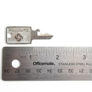 Puede incluir: Una llave plateada con la palabra "Samsonite" impresa en ella. La llave mide 3,5 cm de largo y se mide con una regla.