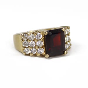 May include: A gold-toned ring featuring a large, square, deep red gemstone at the center. The ring is flanked by clusters of small, round, clear gemstones. The band and setting are a bright, polished gold.