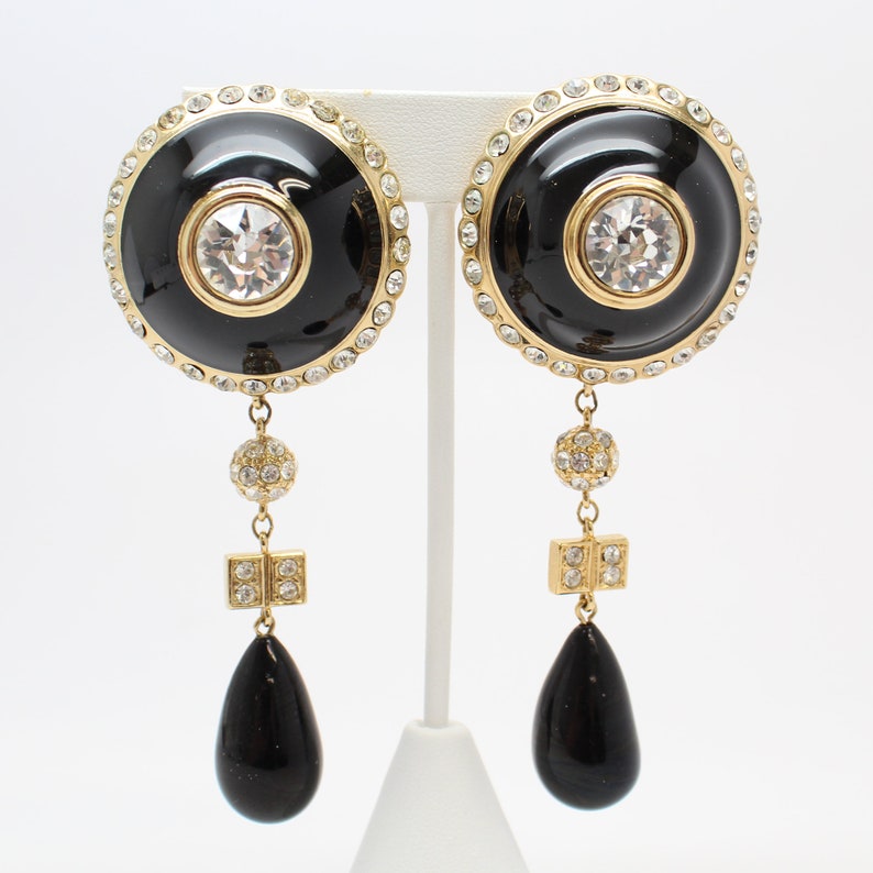 May include: A pair of gold tone dangle earrings with black enamel and clear crystal accents. The earrings feature a round disc with a smaller circle in the center, a crystal ball, a square crystal cluster, and a teardrop-shaped black stone.