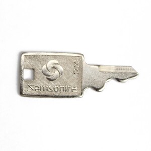 Puede incluir: Una llave plateada con un ojo de cerradura plateado y el texto "Samsonite" y "S0021" en la parte superior. La llave tiene un logotipo circular con tres c&iacute;rculos.