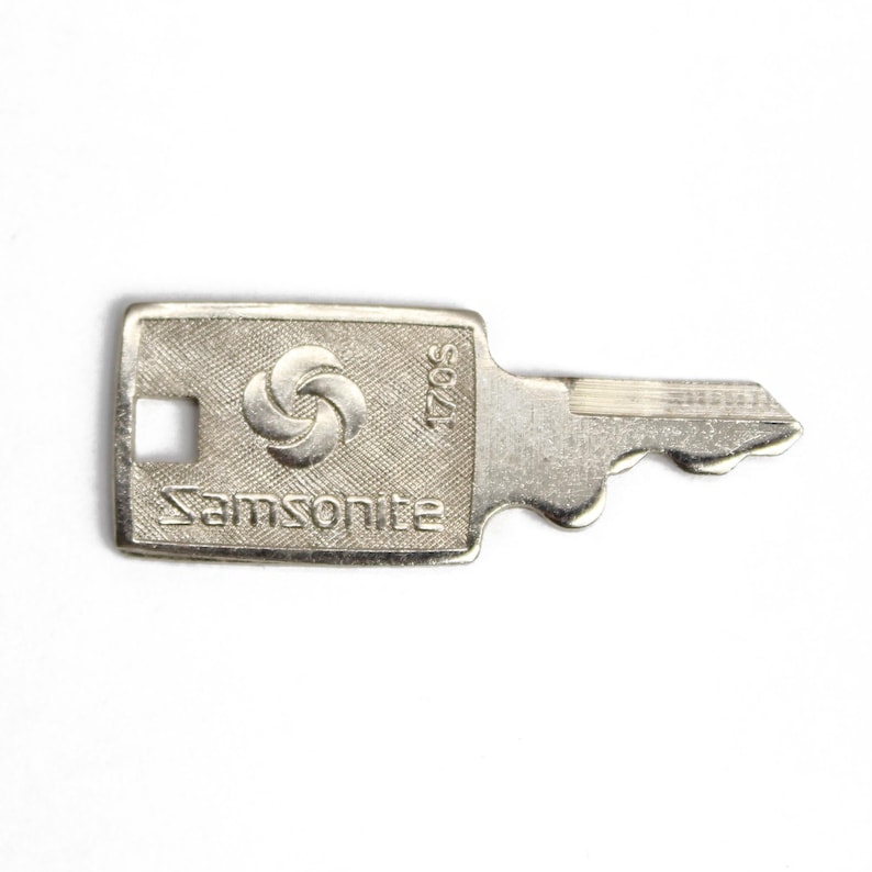 Puede incluir: Una llave plateada con un ojo de cerradura plateado y el texto "Samsonite" y "S0021" en la parte superior. La llave tiene un logotipo circular con tres c&iacute;rculos.