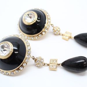 May include: A pair of gold tone dangle earrings with black enamel and clear crystal accents. The earrings feature a round black enamel disc with a clear crystal in the center, surrounded by a row of smaller clear crystals. The disc is attached to a gold tone chain with a black teardrop-shaped pendant.
