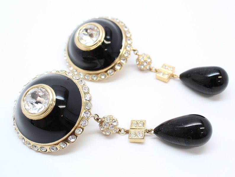 May include: A pair of gold tone dangle earrings with black enamel and clear crystal accents. The earrings feature a round black enamel disc with a clear crystal in the center, surrounded by a row of smaller clear crystals. The disc is attached to a gold tone chain with a black teardrop-shaped pendant.