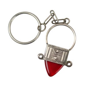 May include: Silver-toned metal keychain with a geometric design and a red, triangular accent. The keychain features a large ring, a decorative rectangular piece with etched patterns, and a smaller, red, teardrop-shaped element.