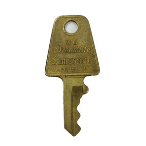May include: A vintage brass key with the text "96 Streamlite Samsonite MADE IN U.S.A." The key has a rectangular head with a circular hole and a notched blade. The key is a golden color.