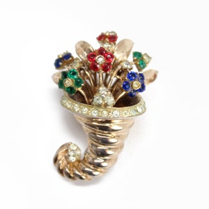 May include: A gold tone brooch with a cornucopia design. The cornucopia is filled with a bouquet of colorful flowers made of rhinestones. The flowers are red, blue, green, and white.