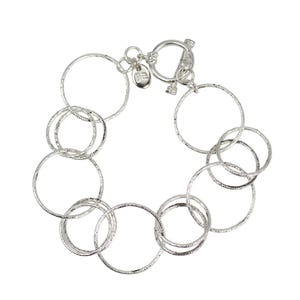 Pode incluir: Uma pulseira prateada com uma série de círculos texturizados interligados. A pulseira tem um fecho de alternância e um pequeno berloque. Os círculos variam em tamanho e estão dispostos em um padrão fluido. A joia está em um fundo branco.