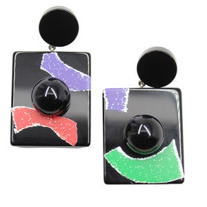 Puede incluir: Un par de pendientes geométricos negros. Cada pendiente presenta una base cuadrada con una esfera negra y acentos coloridos en rojo, morado y verde. La letra "A" es visible en la esfera.
