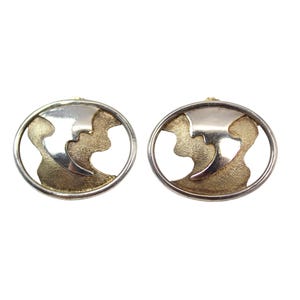 May include: A pair of oval-shaped earrings with a silver-colored border and a gold-colored interior design. The earrings feature a cutout design, creating a unique and modern look. The earrings are displayed against a white background.
