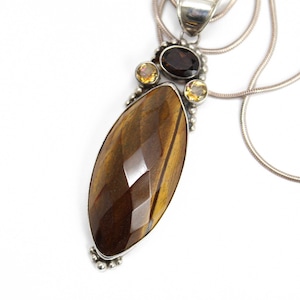 May include: A silver pendant necklace with a large, faceted tiger's eye gemstone. The pendant is accented with smaller, round citrine and smoky quartz gemstones.