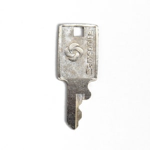 Puede incluir: Una llave plateada con una cabeza rectangular que presenta el logotipo de Samsonite y un dise&ntilde;o circular. La llave tiene un borde dentado.