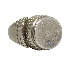 Puede incluir: Anillo plateado con diseño de moneda. El anillo tiene una banda texturizada y un centro circular con detalles intrincados. La moneda parece ser una pieza vintage, agregando una estética antigua y única a la joya.