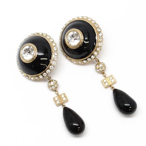 May include: A pair of gold tone drop earrings with black enamel and clear crystal accents. The earrings feature a round, black enamel disc with a clear crystal in the center, surrounded by a row of smaller crystals. The disc is attached to a gold tone chain with a black teardrop pendant.