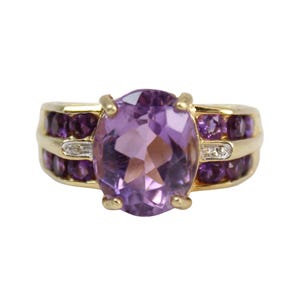 May include: A gold ring featuring a large oval-cut amethyst gemstone as the centerpiece. The ring is accented with smaller, round purple gemstones and two small diamonds. The band is a polished gold color.
