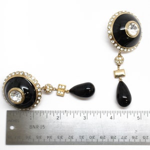 May include: A pair of black and gold dangle earrings with a large black dome-shaped center piece, surrounded by clear rhinestones. Each earring has a smaller black teardrop-shaped dangle and a square-shaped dangle with clear rhinestones.