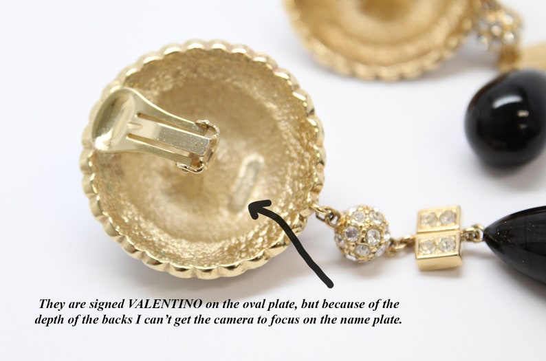 May include: Gold tone clip-on earrings with a black stone and crystal accents. The back of the earring is marked "VALENTINO".