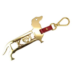 May include: Gold-coloured dachshund-shaped keyring with a red leather strap and a gold clasp. The keyring features the letters "K O I" with a paw print heart design. The brand name "PRADA" is visible on the side.