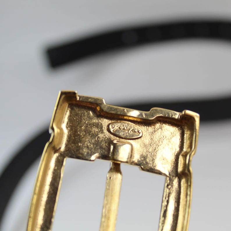 K&ouml;nnte beinhalten: Nahaufnahme einer goldfarbenen Metallschnalle. Die Schnalle hat eine rechteckige Form mit abgerundeten Kanten und einem zentralen Stift. Die Oberfl&auml;che weist eine strukturierte Oberfl&auml;che und eine kleine, ovale Markierung auf. Ein schwarzer G&uuml;rtel ist im Hintergrund zu sehen.